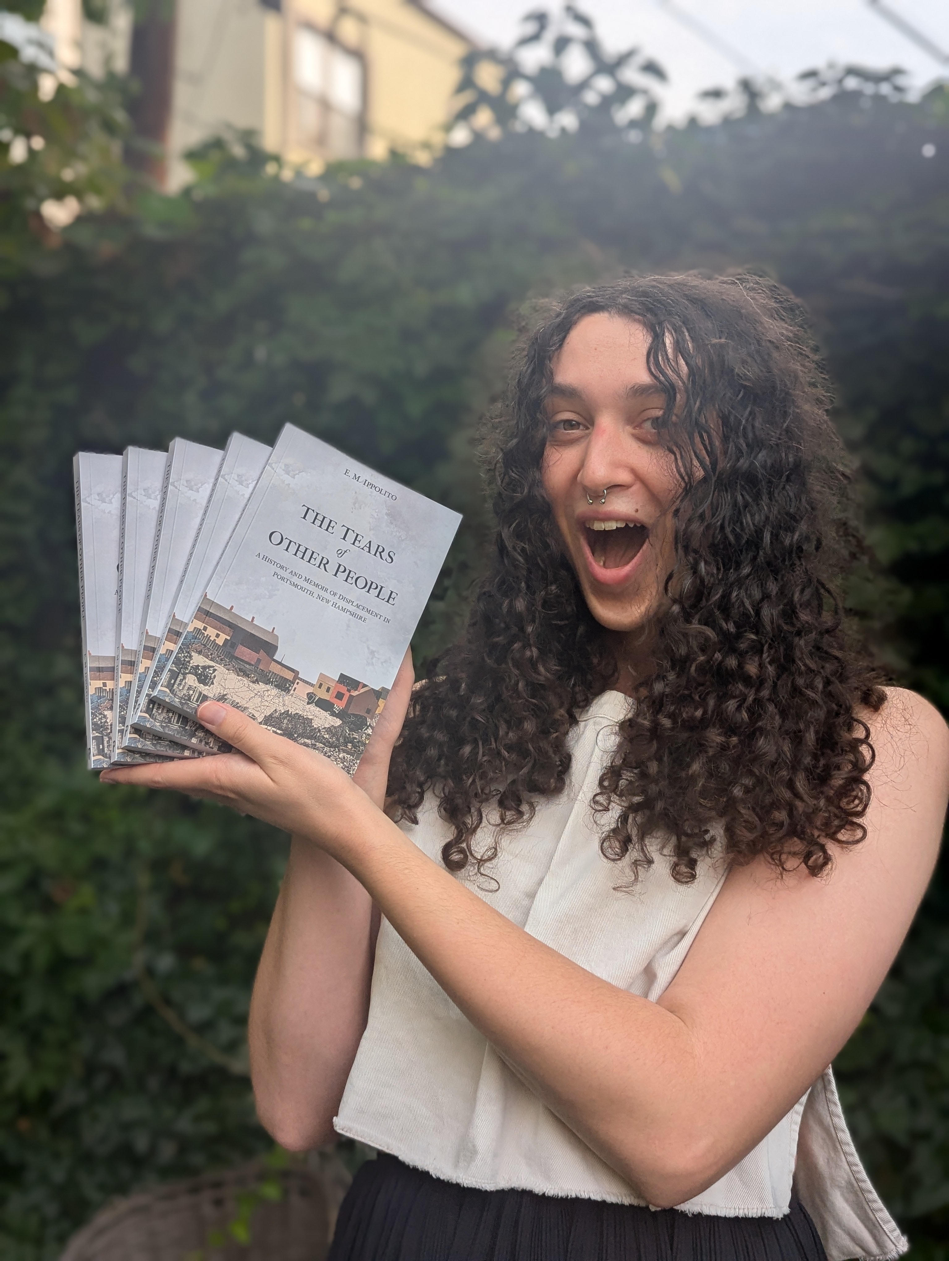 a picture of Evie, a white woman with long curly dark brown hair wearing a sleeveless white shirt and black pleated skirt before a backdrop of green vines, smiling with a nose ring and holding up five copies of the book The Tears of Other People by E. M. Ippolito