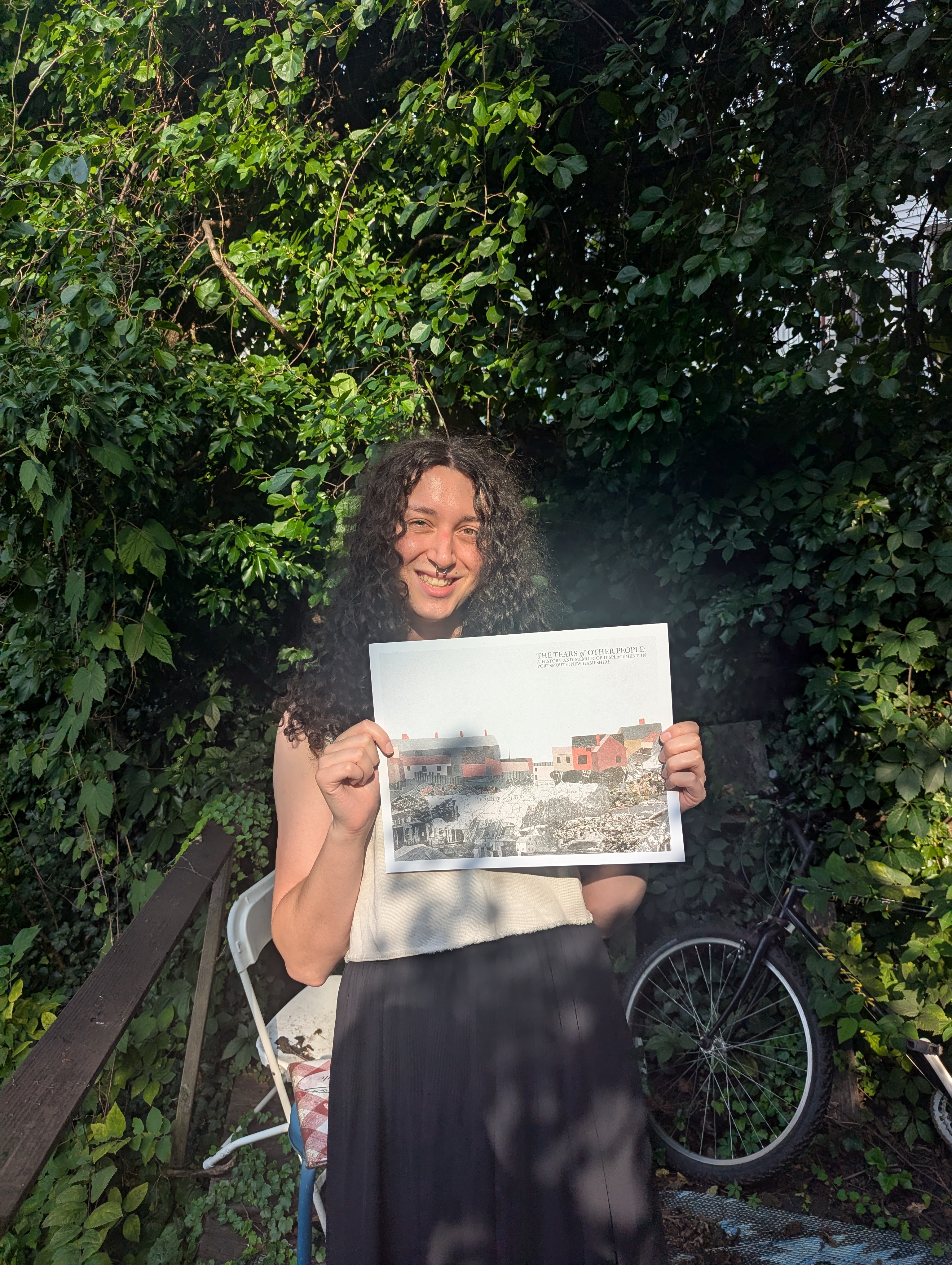 a picture of Evie, same description as previous photo, this time with a patio and a bike in the background, standing in the sun with a poster of the Tears of Other People front cover art by artist Julia Adler @exstrangers